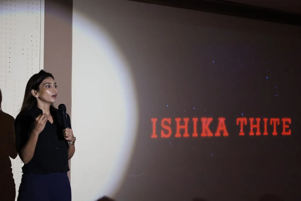 A speaker mid-talk with their title slide projected behind them